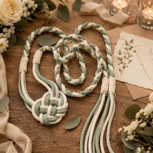 May include: A decorative curtain tieback featuring a braided cord design in sage green and cream white. The tieback includes a large, intricate woven knot at one end and long, loose hanging tassels at the other, secured by wrapped cord accents.