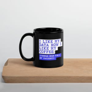 Könnte beinhalten: Ein schwarzer Keramikbecher mit weißem und blauem Text, der lautet: I LIKE MY DATA HOW I LIKE MY COFFEE: STRONG AND FULL OF INSIGHTS. Der Becher hat einen Standardhenkel und einen zylindrischen Körper, wobei die Aufschrift deutlich vorne zu sehen ist.