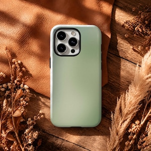 May include: A light sage green smartphone case with a smooth matte finish, featuring a rectangular camera cutout with three circular lenses and a flash, designed to fit a modern mobile device with rounded corners and precise edge cutouts.