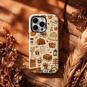 May include: A cream-coloured smartphone case featuring a repeating autumn-themed pattern. The design includes illustrations of pumpkins, a camera, a jumper, books, a basket of apples, a lantern, a bicycle, a mug, and a to-do list on a light background.