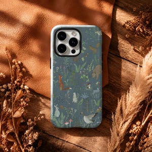 May include: A smartphone case with a slate blue background featuring a repeating pattern of forest animals including a fox, a bear, a rabbit, and a goose, interspersed with various green ferns, wildflowers, and small red mushrooms.