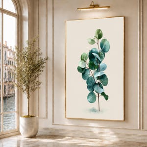 May include: A vertical rectangular wall art print featuring a single eucalyptus branch with rounded, overlapping leaves in shades of deep forest green, teal, and sage, set against a solid, light cream-colored background within a thin gold frame.