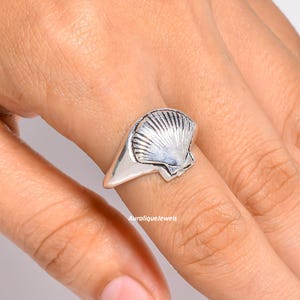 Könnte beinhalten: Ein silberner Ring mit einem detaillierten, fächerförmigen Muscheldesign auf der Vorderseite. Das Metall hat eine polierte Oberfläche mit sichtbaren Rillen, die von der Basis der Muschel ausgehen. Der Text AuraliqueJewels steht in kleinen Buchstaben.