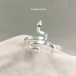 Op de afbeelding: Een zilverkleurige ring in de vorm van een opgerolde slang met een gladde, gepolijste afwerking. Het ontwerp heeft een slangenkop aan de bovenkant en een gebogen staart aan de onderkant, wat een open ring vormt. De tekst AuraliqueJewels is zichtbaar.