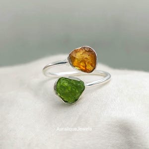 Puede incluir: Un anillo abierto con una banda de plata pulida y dos piedras preciosas en bruto sin tallar. Una piedra es de color ámbar anaranjado translúcido y la otra es de un verde lima vibrante y texturizado. El texto AuraliqueJewels es visible abajo.