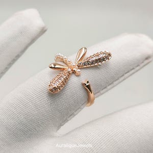 Pode incluir: Um anel aberto em tom de ouro rosa com um design de libélula. A libélula tem duas asas incrustadas com pedras pavé transparentes e duas asas lisas de ouro rosa. O corpo é acentuado com pequenas pérolas brancas e o texto AuraliqueJewels.