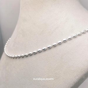 May include: A silver-toned chain necklace composed of small, uniform, oval-shaped beads linked together in a continuous strand. The metal surface has a polished, reflective finish. The text AuraliqueJewels is printed in white at the bottom centre.