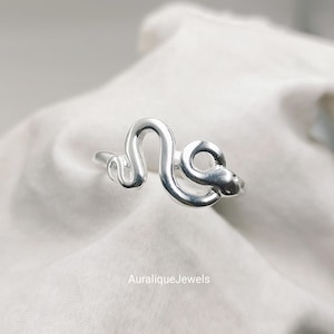 Könnte beinhalten: Ein Ring aus poliertem Silber in Form einer gewundenen Schlange mit einer glatten, reflektierenden Oberfläche. Das Design besteht aus einem durchgehenden Metallband, das den Körper der Schlange bildet, wobei der Kopf auf einer Seite deutlich erkennbar ist. Der Text AuraliqueJewels ist sichtbar.