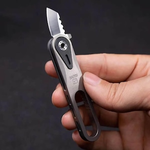 以下が含まれることがあります： メタリックグレーのハンドルと一部が鋸歯状になったシルバーの刃を持つ、コンパクトな折りたたみ式ポケットナイフ。ハンドルには黒いピボットアクセント、ベース部分のカットアウトループ、フレーム側面に刻印された「RSGVE」の文字が特徴です。