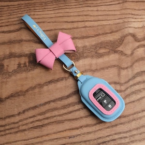 Può includere: Una custodia in pelle azzurra per chiave auto con un fiocco in pelle rosa e un cinturino abbinato. Il cinturino presenta il testo dorato "Tường Vy" e un piccolo cuore dorato. La custodia protegge una chiave auto nera con pulsanti visibili.