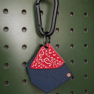 May include: A navy blue faux leather pocket-shaped keychain featuring a red bandana-print fabric insert with white paisley patterns. The pocket is accented with a single copper-colored metal rivet and is attached to a black metal carabiner clip.