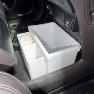 May include: A white plastic multi-compartment storage bin designed for vehicle interiors, featuring a large rectangular main section and a smaller side compartment containing a small Vanicream product container, positioned on the floor.
