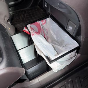 May include: A black plastic car trash bin with a rectangular frame and a divided storage compartment. The bin is lined with a white plastic bag featuring red circular patterns and is positioned in the center console area of a vehicle interior.