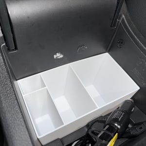 May include: A white plastic center console organizer insert with three distinct compartments of varying sizes, positioned inside a black vehicle storage area next to a black car charger with a visible yellow accent on its plug component.