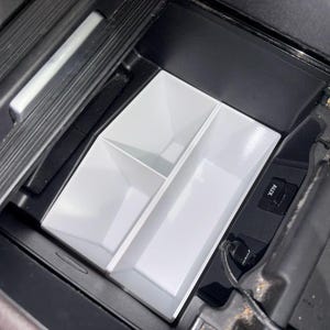 May include: A white plastic center console organizer insert with three distinct storage compartments, designed to fit into a black automotive interior storage space. The insert features a rectangular section and two smaller square sections.