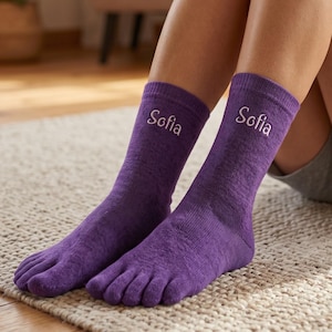 Könnte beinhalten: Ein Paar violette Zehensocken mit einzelnen Fächern für jeden Zeh. Die Socken sind wadenhoch und tragen den Namen Sofia, der in weißer Schreibschrift auf der Vorderseite jeder Socke oberhalb des Knöchelbereichs eingestickt ist.