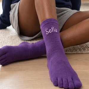 Könnte beinhalten: Ein Paar violette Zehensocken mit einzelnen Fächern für jeden Zeh. Die Socken bestehen aus Strickstoff und tragen den Namen Sofia, der in weißer Schreibschrift auf der Außenseite des Knöchelbereichs der linken Socke eingestickt ist.