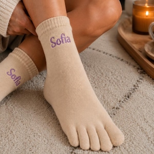 Könnte beinhalten: Ein Paar beigefarbene Zehensocken mit dem in violettem Garn gestickten Namen Sofia am gerippten Bündchen. Die Socken haben einzelne Fächer für jeden Zeh und eine wadenlange Passform, gefertigt aus einem weichen Strickmaterial.