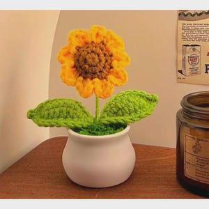 May include: A crocheted sunflower with bright yellow petals and a brown center, featuring two green leaves on a thin green stem. The flower is planted in a small, smooth, white ceramic pot with a rounded base, creating a decorative display.