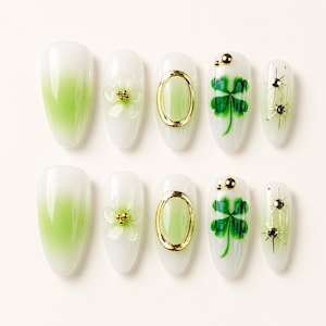 May include: A set of ten almond-shaped press-on nails featuring a white and light green gradient base. Designs include a 3D white flower with gold beads, a gold oval frame, a green four-leaf clover with gold studs, and gold starburst accents.