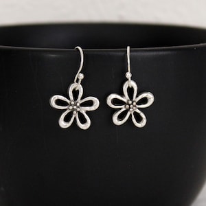 May include: A pair of silver-colored flower earrings with a hook closure. Each earring features a five-petal flower design with a small, textured center. The earrings are displayed against a black background.