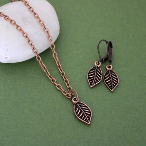 May include: A copper-toned jewelry set featuring a leaf pendant necklace and matching leaf earrings. The necklace has a delicate chain, and the earrings have lever-back closures. The leaf pendants and earrings have detailed vein patterns.