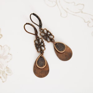 May include: A pair of copper-toned earrings with a flower design and a teardrop shape. The earrings have a hook closure.