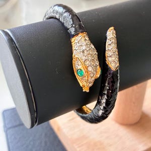 以下が含まれることがあります： 黒い質感のあるボディ、クリアなラインストーンが散りばめられたゴールドトーンの頭部、そして緑色の石の目が特徴の、巻き付くタイプのスネークブレスレット。尻尾の先端もゴールドトーンの金属とクリアなラインストーンで仕上げられています。