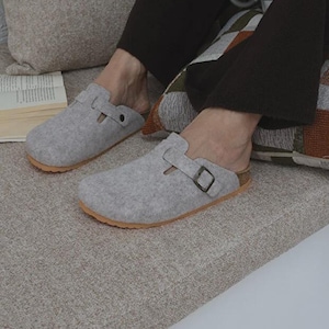 May include: A pair of light gray felt clogs featuring a single strap across the top with an adjustable dark metal buckle. The footwear has a rounded toe design and a tan-colored sole, providing a slip-on style with an open back construction.
