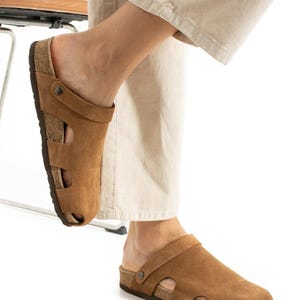 May include: A pair of tan suede clogs featuring a closed-toe design with side cutouts, a wide strap across the instep secured by a metal rivet, and a contoured cork footbed with a dark brown rubber outsole for traction and support.