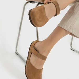 May include: A pair of tan suede shoes featuring a rounded toe, a closed heel, and a single adjustable strap across the instep with a hook-and-loop closure. The shoes have a dark brown sole and a textured, matte finish on the exterior suede material.
