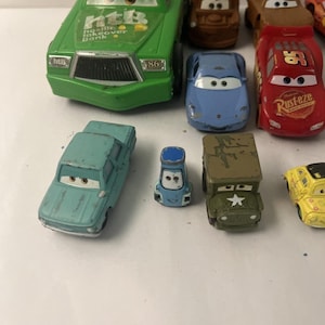 Puede incluir: Una colecci&oacute;n de coches de juguete de metal que representan varios personajes de la franquicia Cars. El conjunto incluye un coche de carreras verde grande con el texto 'htB', un coche de carreras rojo con '95' y 'Rust-eze', un coche deportivo azul, una gr&uacute;a marr&oacute;n, un sed&aacute;n azul claro, un veh&iacute;culo azul peque&ntilde;o, un jeep militar verde y un coche amarillo.