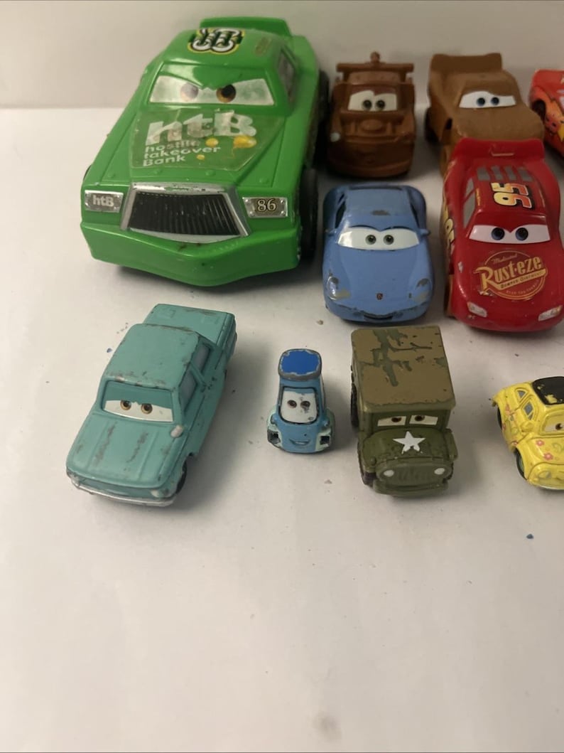 Puede incluir: Una colecci&oacute;n de coches de juguete de metal que representan varios personajes de la franquicia Cars. El conjunto incluye un coche de carreras verde grande con el texto 'htB', un coche de carreras rojo con '95' y 'Rust-eze', un coche deportivo azul, una gr&uacute;a marr&oacute;n, un sed&aacute;n azul claro, un veh&iacute;culo azul peque&ntilde;o, un jeep militar verde y un coche amarillo.