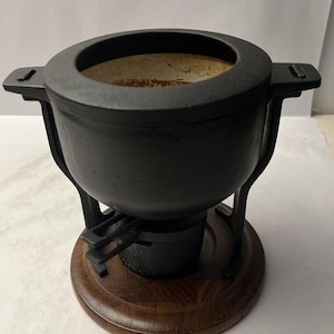 Puede incluir: Una olla para fondue de hierro fundido negro con una abertura circular y dos asas laterales, apoyada sobre un soporte de metal negro. La olla se asienta sobre una base redonda de madera marrón oscuro con veta visible. El interior es claro.
