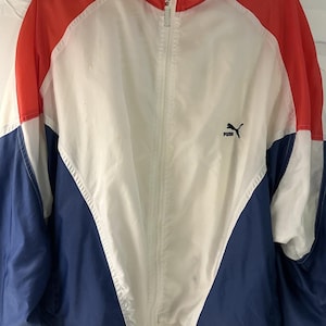 Op de afbeelding: Een Puma trainingsjack met ritssluiting en een kleurblokontwerp in rood, wit en blauw. Het jack heeft een hoge kraag, een volledige ritssluiting aan de voorzijde en het zwarte Puma-logo en de tekst geborduurd op de linkerborst.