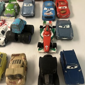 May include: A collection of die-cast toy cars featuring various models, including a red race car with the number 95, a blue Dinoco car, a green military vehicle, a formula-style car with Italian flag colors, and several other colorful character vehicles.