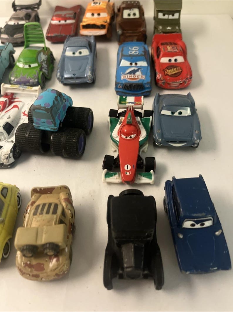 May include: A collection of die-cast toy cars featuring various models, including a red race car with the number 95, a blue Dinoco car, a green military vehicle, a formula-style car with Italian flag colors, and several other colorful character vehicles.
