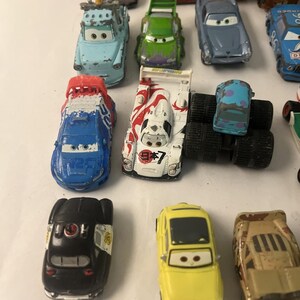 May include: A collection of various die-cast toy cars featuring diverse colors, shapes, and character designs. The assortment includes a blue race car, a white car with red Japanese text, a black police car, a yellow car, and several other vehicles.