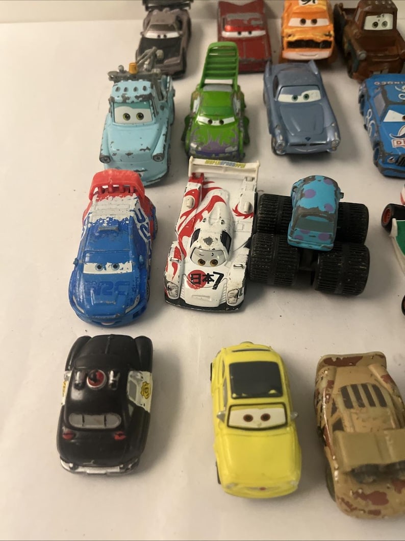 May include: A collection of various die-cast toy cars featuring diverse colors, shapes, and character designs. The assortment includes a blue race car, a white car with red Japanese text, a black police car, a yellow car, and several other vehicles.