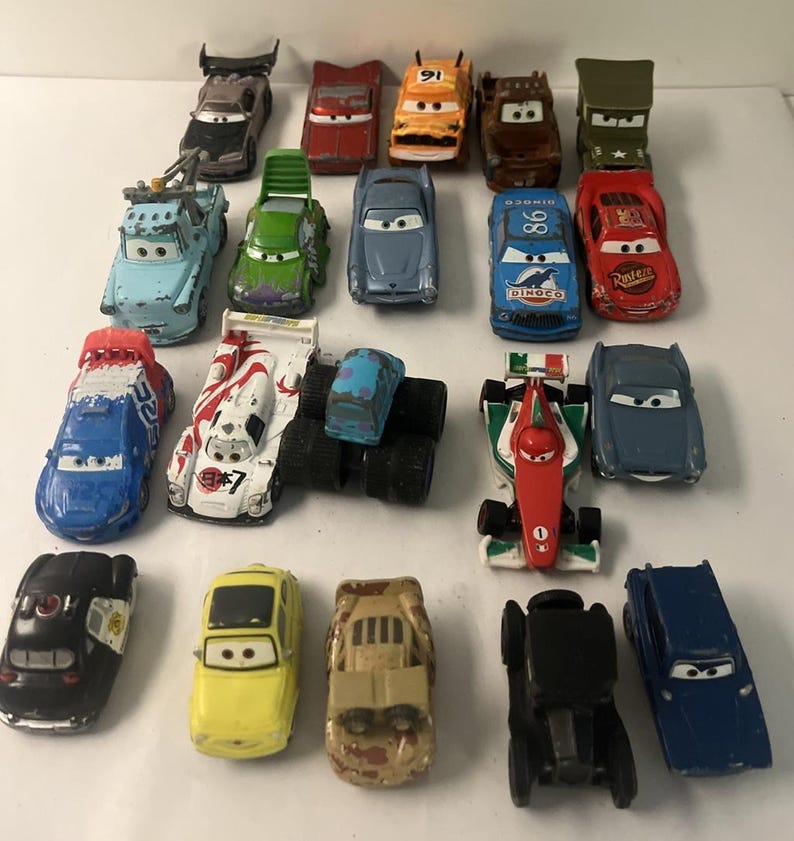 May include: A collection of nineteen die-cast toy cars featuring various colors, shapes, and character designs. The assortment includes race cars, trucks, and vintage vehicles, each with painted details, windshield eyes, and unique body styles.