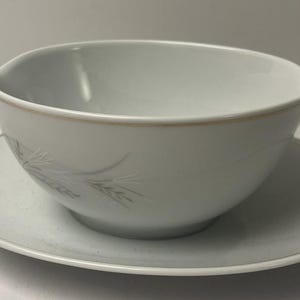 May include: A white porcelain gravy boat with a spout and a matching saucer. The bowl features a delicate, light grey wheat stalk pattern and a thin gold rim along the top edge of the bowl and the outer edge of the saucer, creating a classic look.