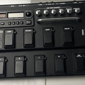 Puede incluir: Un procesador de efectos para guitarra Line 6 POD XT Live de color negro con pantalla digital, múltiples interruptores de pie, perillas de control de tono de amplificador y un pedal de expresión integrado, con etiquetas Variax, Bank, Amp, Stomp y Mod.