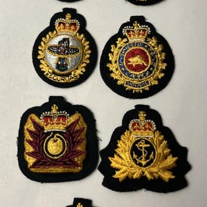 May include: Seven embroidered military-style patches on a black fabric base. Each patch features gold thread detailing, a crown emblem at the top, and unique central symbols including an anchor, a beaver, a totem pole, and various geometric shapes.
