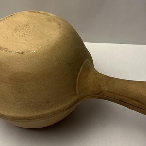 May include: A tan, round, unglazed stoneware pan with a single handle extending from the side. The surface has a matte, earthy texture with a circular stamp visible on the top centre. The design features a rounded bowl shape with a distinct rim.