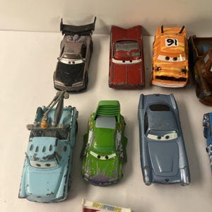 May include: A collection of die-cast toy cars featuring various models including a grey car with a spoiler, a red car, an orange car with the number 91, a light blue tow truck, a green car with purple accents, and a metallic blue sports car.