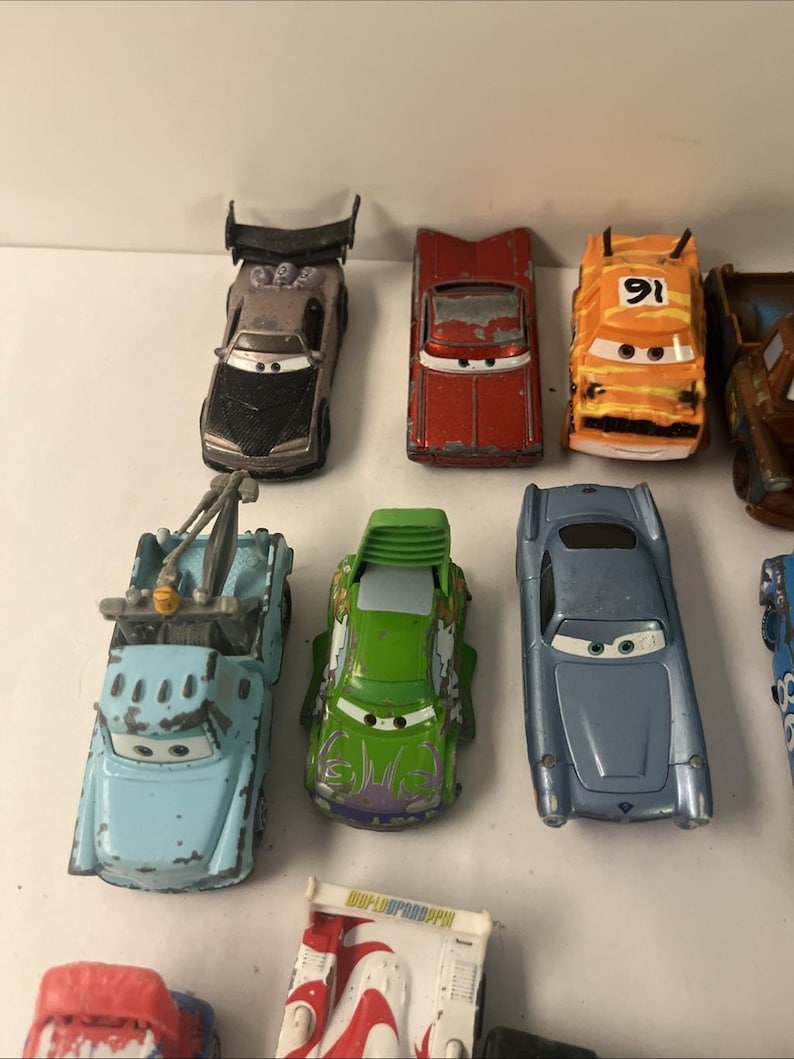 May include: A collection of die-cast toy cars featuring various models including a grey car with a spoiler, a red car, an orange car with the number 91, a light blue tow truck, a green car with purple accents, and a metallic blue sports car.