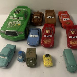 Puede incluir: Una colecci&oacute;n de once coches de juguete de metal que representan varios personajes de la franquicia Cars, incluyendo un coche verde grande de Hostile Takeover Bank, corredores rojos de Lightning McQueen, una gr&uacute;a marr&oacute;n y otros veh&iacute;culos.