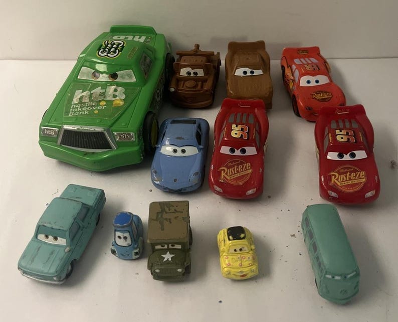 Puede incluir: Una colecci&oacute;n de once coches de juguete de metal que representan varios personajes de la franquicia Cars, incluyendo un coche verde grande de Hostile Takeover Bank, corredores rojos de Lightning McQueen, una gr&uacute;a marr&oacute;n y otros veh&iacute;culos.