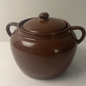 May include: A round, dark brown ceramic pot with a glossy finish, featuring two curved side handles and a matching removable lid with a small knob handle on top. The vessel has a wide, bulbous body and a smooth, uniform glazed surface.