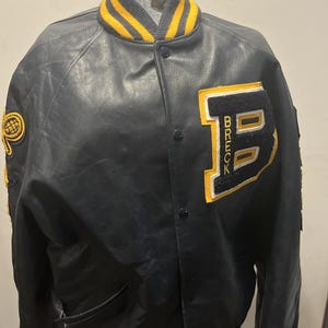 May include: A dark navy blue varsity jacket featuring a large felt letter B with the word BRECK embroidered inside. The jacket has a snap-button front, a ribbed collar with yellow stripes, and a yellow and black embroidered tennis racket patch.